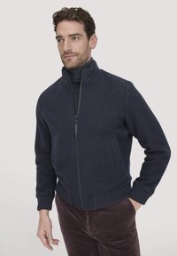 Man in een marineblauwe ritsjack over een donkere turtleneck, met bruine corduroy broek, staande tegen een effen lichte achtergrond.