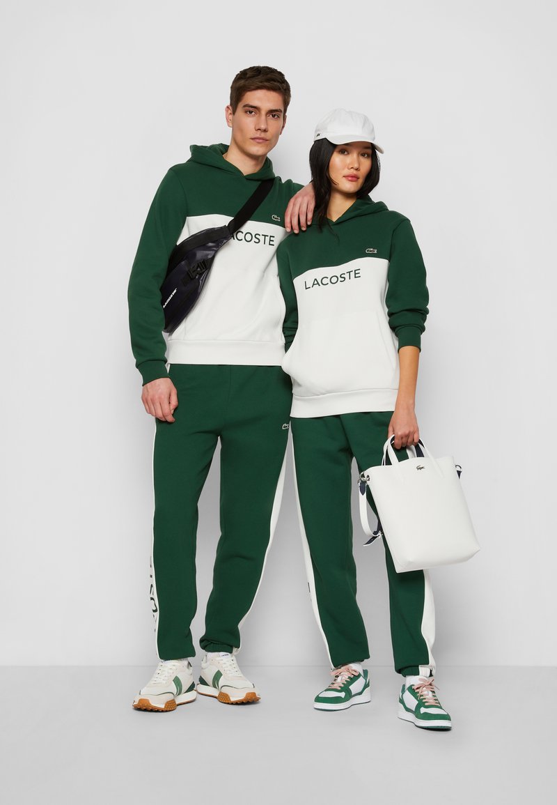 Uomo e donna che indossano felpe e pantaloni della tuta verdi e bianchi di Lacoste, scarpe bianche, uomo con borsa a tracolla nera, donna con cappellino bianco e borsa bianca.