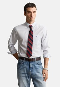 Chemise blanche boutonnée avec manches retroussées, cravate rayée bleu marine et rouge, jean bleu, ceinture marron, montre argentée. Design simple et classique.