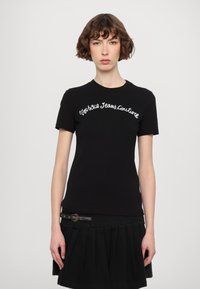Melna pieguļoša T-krekls ar baltu izšūtu "Versace Jeans Couture" tekstu priekšā, īsām piedurknēm un apaļu kakla izgriezumu.