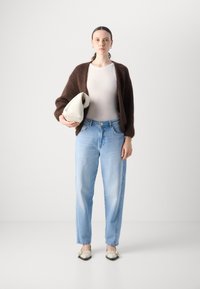 Brauner, weicher Cardigan, weißes, figurbetontes Oberteil, hellblaue gerade Jeans, hält eine beigefarbene Tasche und trägt flache Schuhe mit schwarzer Verzierung.