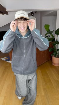 Grå fleecejakke med lynlås og lyseblå detaljer, grå joggingbukser, klare briller og en lys beige kasket med sort tekst. Indendørs miljø.