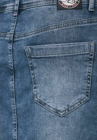 Blå denimjeans med en bakficka, synlig sömnad och en märkesetikett vid midjan. Texturen ser slät ut.