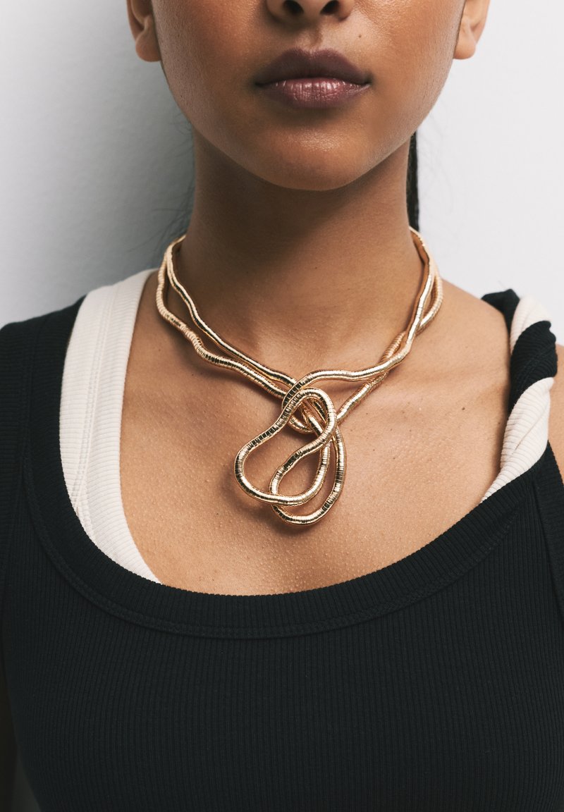 Collana in metallo color oro con fili intrecciati che formano un design a nodo. Presenta una texture liscia e lucida. Indossata sopra una maglietta nera.