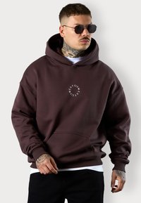 Sweat à capuche marron avec une poche avant et un logo circulaire blanc. Il est doté d'une capuche spacieuse et de poignets côtelés, fabriqué à partir d'un tissu doux.