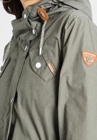 Oliwkowa kurtka z kapturem, wyposażona w srebrne guziki, klapy kieszeni z przodu oraz teksturowany materiał. Posiada brązową skórzaną naszywkę z logo oraz sznurek do regulacji.