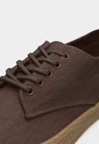 Brązowy materiałowy sneaker z dopasowanymi sznurowadłami, metalowymi oczkami i beżową, teksturowaną gumową podeszwą, pokazany z bliska pod kątem.