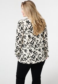 Chemisier à manches longues en tissu crème avec un motif floral noir. Le design présente une finition texturée et une coupe décontractée. Associé à un pantalon noir.