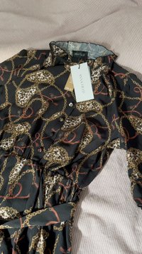 Vestido negro con cadena y diseño de leopardo. Presenta un cuello con solapa, botones en la parte delantera y cintura fruncida. Incluye etiqueta de la marca.