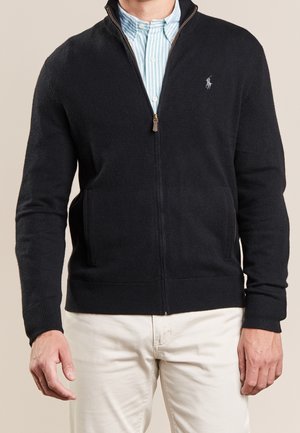 Uomo con maglione nero con zip sopra una camicia a righe blu e pantaloni bianchi, in piedi davanti a uno sfondo beige.