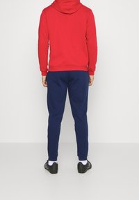 Sudadera roja con capucha y pantalones de chándal azul marino. La sudadera tiene un bolsillo tipo canguro; los pantalones de chándal tienen puños elásticos. Se ven zapatillas deportivas negras.