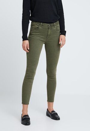 Vrouw draagt olijfgroene skinny broek, zwart shirt met lange mouwen en zwarte loafers, staand tegen een effen achtergrond.