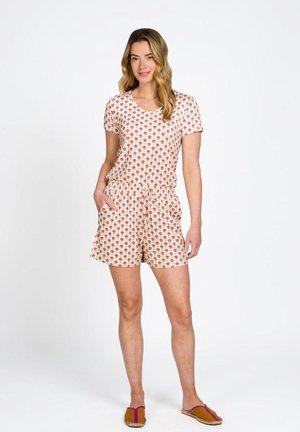 Vrouw staand met één hand in de zak, gekleed in een bijpassende top met korte mouwen en korte broek met een klein rood patroon en bruine instapschoenen.