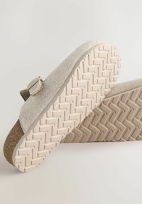 Zuecos de fieltro beige con acento de corcho, correa ajustable y suela de goma texturizada con patrón de espiga para mayor tracción.