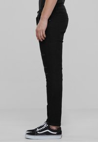 Jeans slim noir avec des accents déchirés, associés à des baskets Vans noir et blanc. La vue de côté met en valeur la texture du tissu et la coupe.