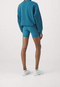 Osoba w turkusowej, oversize'owej bluzie i dopasowanych, obcisłych szortach rowerowych, stojąca tyłem do kamery, w białych sneakersach Adidas na jednolitym tle.