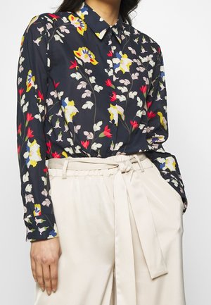 Kvinde iført en mørk blomstret langærmet bluse, stoppet ned i high-waisted beige bukser med et stofbælte bundet i en knude.