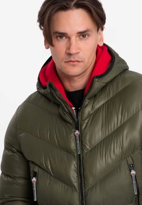 Olivgrön pufferjacka med röd huva, diagonalt chevronmönster, dragkedjefickor och svarta detaljer längs dragkedjorna och kanterna.