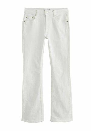 Femme portant un blazer texturé écru sur mesure avec ceinture, un pantalon large assorti et des talons métalliques à bout pointu, debout contre un mur clair.