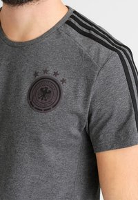 Harmaa lyhythihainen t-paita, jossa on pyöreä musta emblem, jossa on kotka ja viisi tähteä, sekä mustat kolmiraitadetailit olkapäillä.