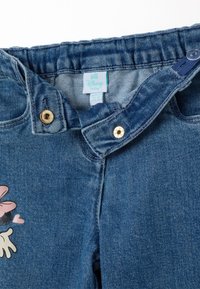 Jeans de bebé de mezclilla con un lavado azul claro, con cierre de botón dorado, cintura elástica y un gráfico de flor rosa en la parte frontal.