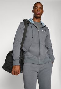 Grijze zip-up hoodie met een lichtblauwe capuchon, gedragen met bijpassende joggers. Het model houdt een zwarte sporttas vast. Merklogo zichtbaar op de borst.