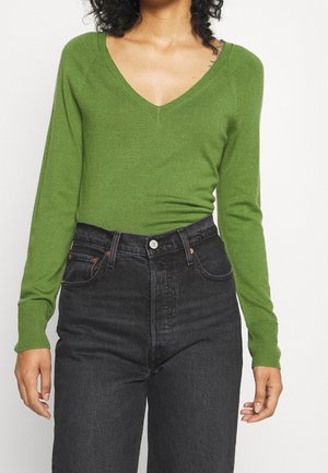 Personne portant un pull vert ajusté à col en V et un jean noir taille haute, montré du cou aux hanches sur un fond uni.