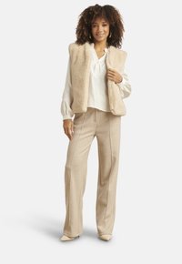 Vrouw draagt een beige faux fur-vest, witte blouse met lange mouwen, beige broek met wijde pijpen en beige puntige schoenen, staand en glimlachend.