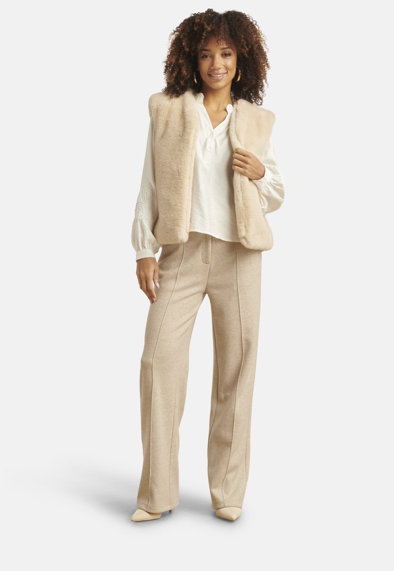 Vrouw draagt een beige faux fur-vest, witte blouse met lange mouwen, beige broek met wijde pijpen en beige puntige schoenen, staand en glimlachend.