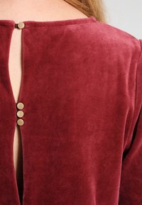 Burgundowa aksamitna bluzka z okrągłym dekoltem i zapięciem na guziki z tyłu. Posiada trzy złote guziki wzdłuż otwarcia.
