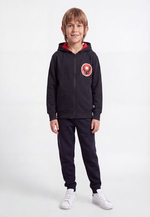 Ung pojke står och ler, bär en svart zip-up hoodie med en röd och svart logga, svarta byxor och vita sneakers.