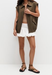 Oliwkowa zielona kamizelka bez rękawów na zamek błyskawiczny, z bocznymi kieszeniami, noszona na beżowym krótkim topie i białych szortach z falbankami; do tego brązowe sandały na płaskiej podeszwie.