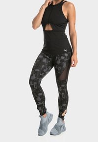 Musta hihaton toppi leikatulla yksityiskohdalla, yhdistettynä istuviin mustiin kuvioituihin leggingsseihin, joissa on verkkopaneelit, ja harmaat urheilukengät.