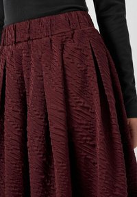 Burgundy plisseret nederdel med en struktureret overflade og elastisk talje. Stoffet viser et bølget mønster, der forbedrer dens dimension.