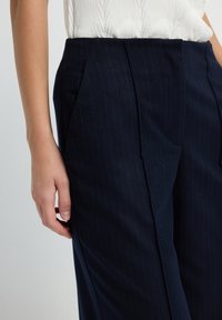 Vrouw in donker marineblauwe pinstripe broek met zijzakken en een witte textuurloze mouwloze top, staand met haar linkerhand ontspannen.