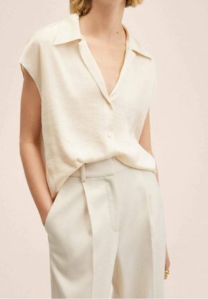 Mouwloze crèmekleurige blouse met kraag en knopen, ingestopt in crèmekleurige broek met hoge taille, linkerhand van het model in de zak.