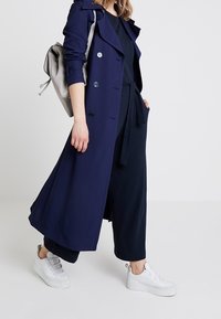 Žena v námořnickém dlouhém kabátu, černých širokých kalhotách, bílých platformových teniskách a s světle šedým batohem na rameni.