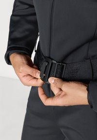 Μαύρη στολή κατάδυσης με υφή υφάσματος, που διαθέτει μια φαρδιά, ρυθμιζόμενη μαύρη ζώνη και ανθεκτικό κούμπωμα, κρατημένη από ένα χέρι που ρυθμίζει την εφαρμογή.