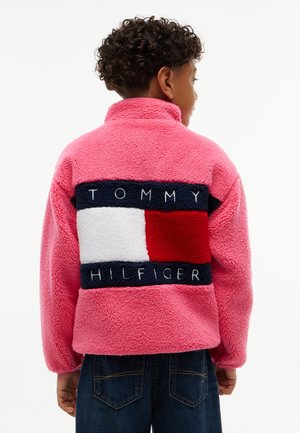 Dziecko z kręconymi włosami, ubrane w jaskraworóżową polarową kurtkę z dużym logo Tommy Hilfiger na plecach oraz ciemnoniebieskie dżinsy.