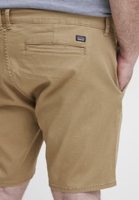 Beige Shorts aus Baumwollstoff, ausgestattet mit einer Gesäßtasche und einem kleinen schwarzen Etikett. Sie haben eine gerade Passform und enden knapp über dem Knie.