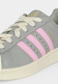 Γκρι αθλητικό παπούτσι Adidas Superstar με ροζ ρίγες, λευκά κορδόνια, χρυσό λογότυπο στη γλώσσα και χρυσό κείμενο "Superstar" στο πλάι.