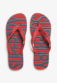 Tongs en caoutchouc rouge avec un marquage gris. Semelle intérieure texturée avec un motif répétitif "SUPERDRY". Bracelets plats et flexibles avec un design en chevrons.