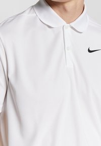 Bílá polo košile z texturované tkaniny, s klasickým límcem, třemi knoflíky a černým logem Nike na levé hrudi.