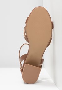 Sandale marron à semelle plate, avec une base beige texturée et des lanières entrecroisées. Elle dispose également d'accents en matériel décoratif.