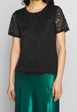 Top negro de encaje de mangas cortas con escote redondo, que presenta patrones florales y un dobladillo festonado. Combinado con una falda de satén verde.