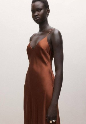 Mujer alta de piel oscura con un vestido sin mangas de raso color óxido y anillos plateados, posando frente a un fondo claro y liso.