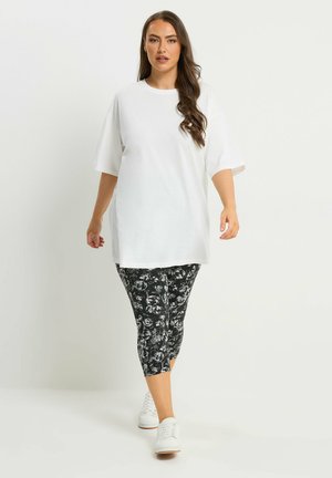 Kvinde går iført en løs, hvid kortærmet skjorte, sort-hvide mønstrede cropped leggings og hvide sneakers.