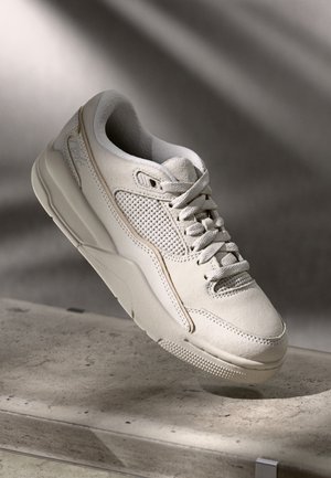 Sneaker w kolorze ecru z panelami ze zamszu i siateczki, grubą podeszwą i sznurowadłami w układzie krzyżowym, ustawiony na teksturowanej kamiennej powierzchni.