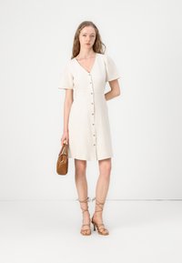 Beige linnen jurk met korte, geplooide mouwen en een diepe V-hals. Heeft een knoopsluiting aan de voorkant en een getailleerde taille. Geribbelde bruine handtas.
