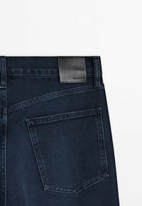 Jeans blu scuro con una toppa in pelle nera che presenta un marchio stampato, una singola tasca posteriore e dettagli di cucitura in stile denim classico.